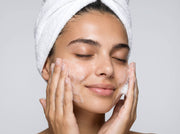 Woman applying cream to her face with a white towel on her head against a light gray background