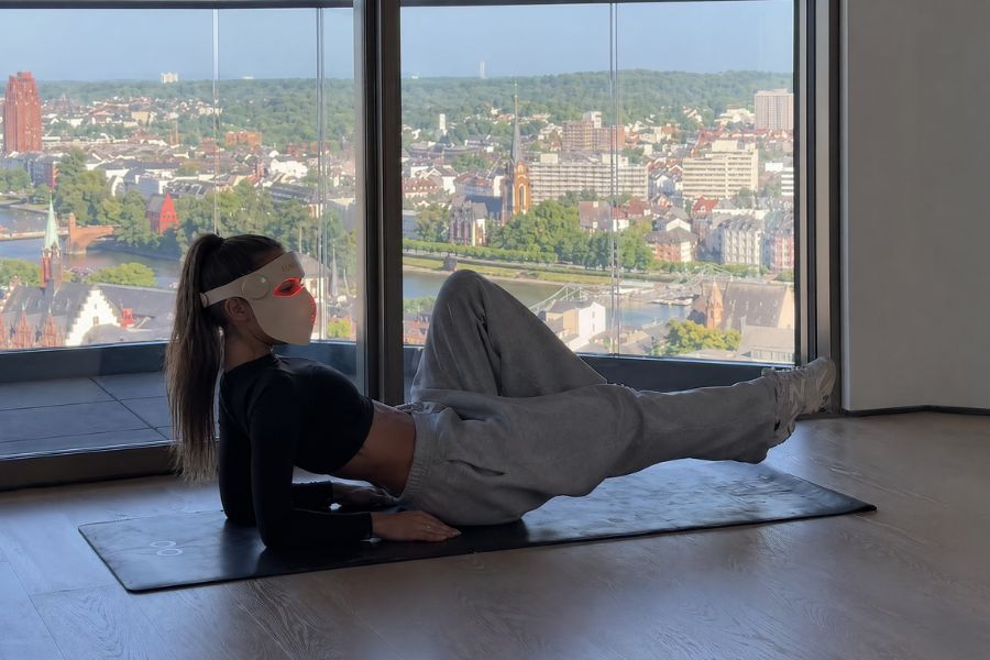 Person exercising with luscent red red light therapy mask on a mat with a cityscape view from large windows.