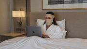 Woman in a robe using a laptop on a bed with a nightstand and lamp in the background.