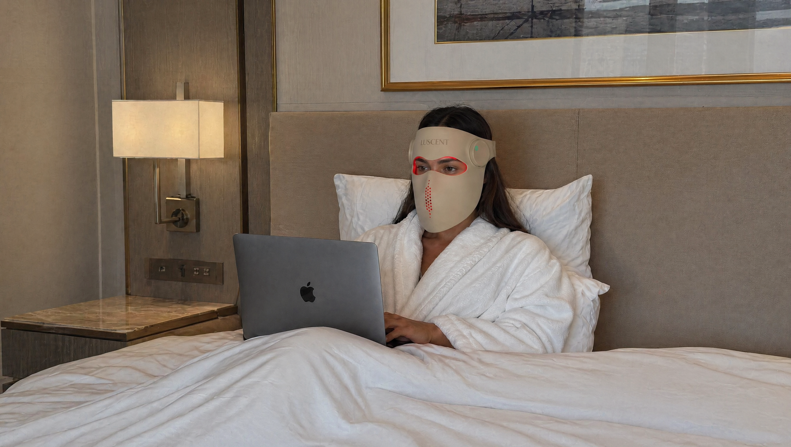 Woman in a robe using a laptop on a bed with a nightstand and lamp in the background.