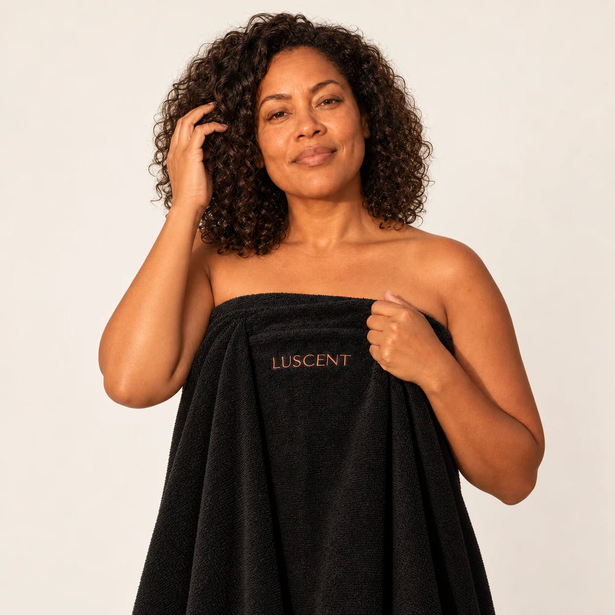 Woman holding a black Luscent sauna blanket insert with one hand near her curls against an ivory studio background